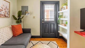 A living room with a dark door, a grey sofa, a red pillow, a floor lamp, and a wall-mounted TV. - Perfect Hawthorne Micro By Top Shopping & Dining (Portland)