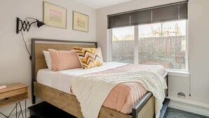 Een slaapkamer met een houten bed, een nachtkastje, een raam met jaloezieën en twee ingelijste foto's aan de muur.