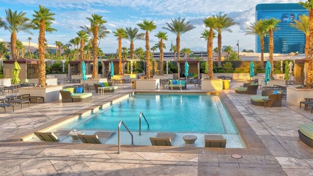 A pool area with lounge chairs and umbrellas, surrounded by palm trees and a modern building in the background. - MGM Signature Balcony One Bedroom Two Beds Suite (Las Vegas)