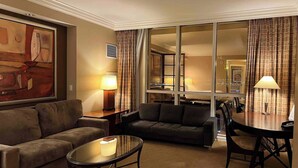 A hotel room with a living area featuring two sofas, a glass coffee table, and a dining table with chairs. There is a large window with curtains, a painting on the wall, and a lamp on a side table. - MGM 1 Bedroom Balcony And Or Studio Luxury Suites (Las Vegas)