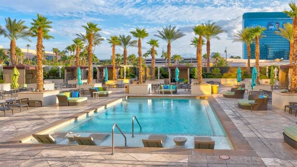 A pool area with lounge chairs and umbrellas, surrounded by palm trees and a modern building in the background. - MGM 1 Bedroom Balcony And Or Studio Luxury Suites (Las Vegas)