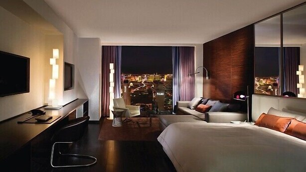 Habitació d'hotel moderna amb un llit gran, una taula de treball, una cadira i un televisor. Es veu la ciutat de nit a través de la finestra.