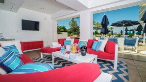 Un espace détente moderne avec des sièges rouges et bleus, une table basse blanche avec des boissons, et une vue sur la piscine extérieure.