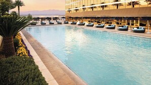 Une grande piscine entourée de chaises longues et de parasols, le tout bordé d’un bâtiment moderne doté de grandes fenêtres.