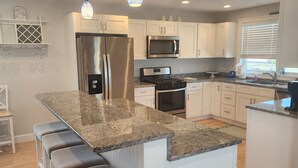 A modern kitchen with white cabinets, stainless steel appliances, a granite countertop, and bar stools. - Spacious Beach Home - Steps to Ocean, Dining & Entertainment (Salisbury)