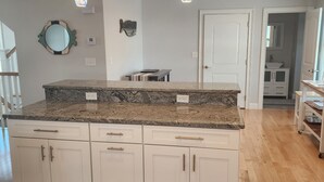 A kitchen with a granite countertop, white cabinets, and a fish-shaped mirror. There are pendant lights, a door, and a hallway leading to another room. - Spacious Beach Home - Steps to Ocean, Dining & Entertainment (Salisbury)