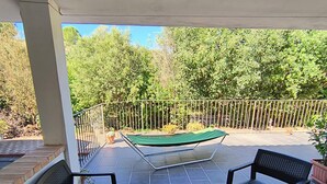 Eine überdachte Terrasse mit zwei schwarzen Metallstühlen und einem kleinen Tisch, einer grünen Hängematte und einem Pool im Hintergrund.