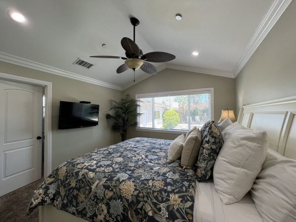 A bedroom with a bed, a ceiling fan, a TV, and a window with a view.