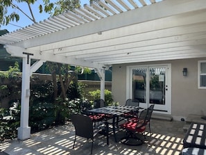 Un patio exterior con una mesa y sillas cubiertas, una pérgola y una puerta que da a un área techada.