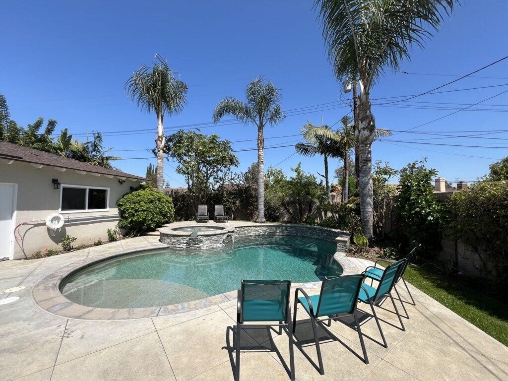 A swimming pool with a curved edge, surrounded by a patio with chairs, palm trees, and a house in the background.