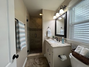 A bathroom with a shower, a sink with a mirror, and a window with blinds.