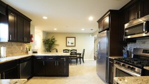 A modern kitchen with dark cabinetry, a central island, stainless steel appliances, and a dining area with chairs and a table. - Awesome 4 Bedroom, 3 1/2 Bath Home 1/2 Mile Walk To Disney (Anaheim)