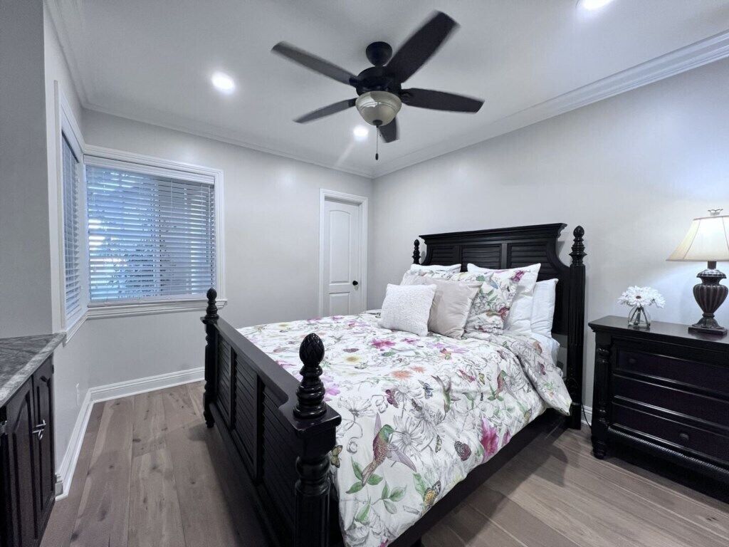 A bedroom with a bed, a ceiling fan, a window with blinds, a dresser, and a lamp.