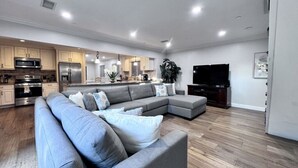 A modern living room with a sectional sofa, a flat-screen TV, and a kitchen in the background. - Spectacular 5 Bedroom, 4 Bath Pool Home Near Disney (Anaheim)