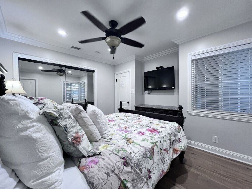 A bedroom with a bed, a ceiling fan, a television, and a window with blinds.