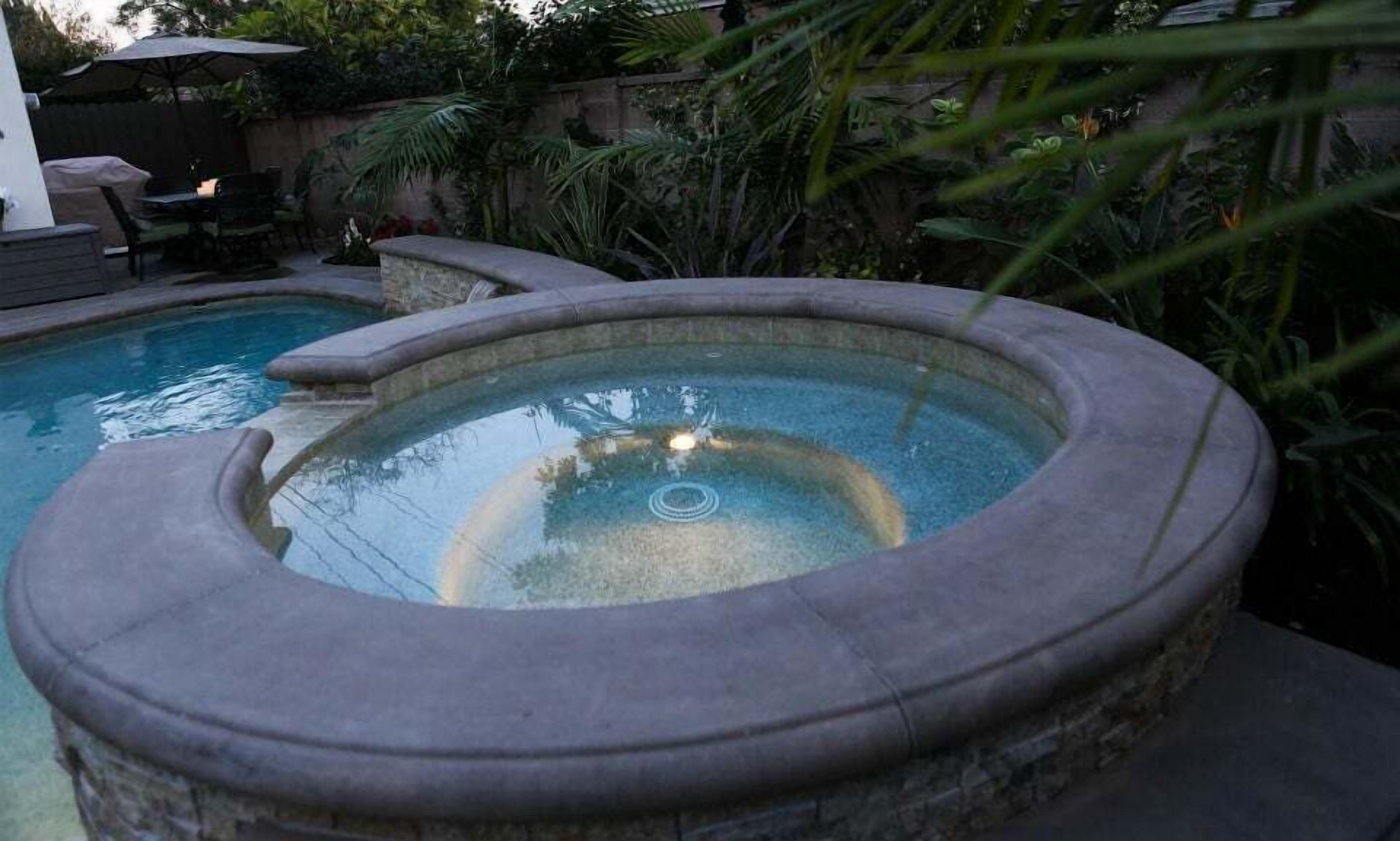 A stone hot tub in a backyard with a pool and outdoor furniture.