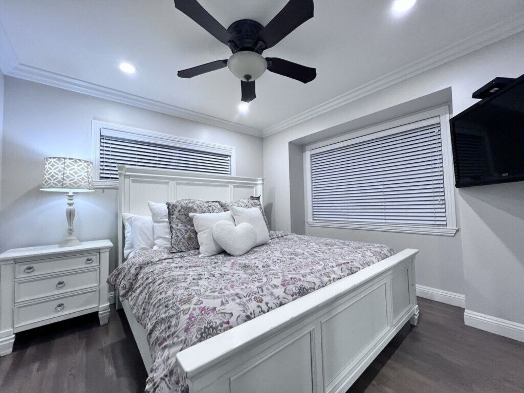 Une chambre à coucher comprenant un lit, une table de chevet, une lampe, un ventilateur de plafond et un téléviseur fixé au mur.