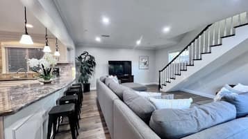 Una sala moderna con un sofá gris, taburetes negros, una isla de cocina y una escalera.