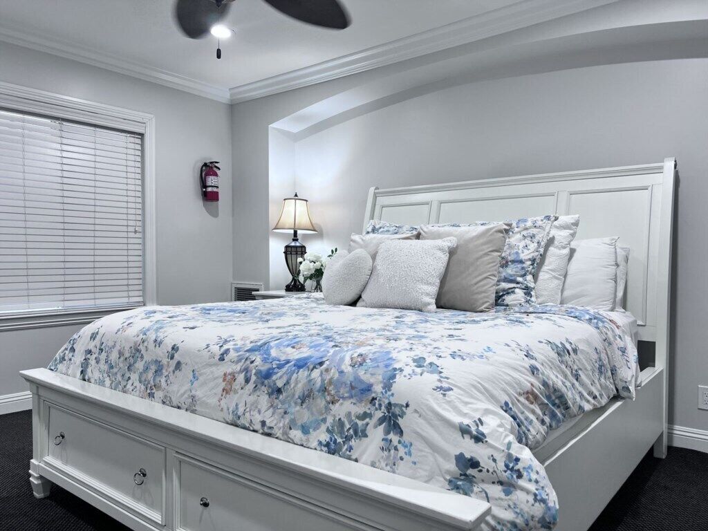 Une chambre à coucher avec un lit blanc, une table de chevet, une lampe et un ventilateur de plafond.