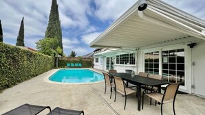 A swimming pool with lounge chairs, a covered patio with a table and chairs, and a house with a white exterior and large windows. - Large Family Pool Home 2.5 Miles To Disney w/3 King Beds!!! (Anaheim)
