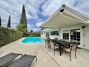 A swimming pool with lounge chairs, a covered patio with a table and chairs, and a house with a white exterior and large windows.