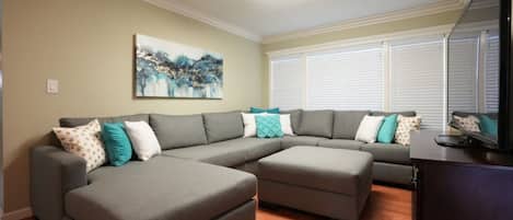 A living room with a sectional sofa, ottoman, and a flat-screen TV on a cabinet.