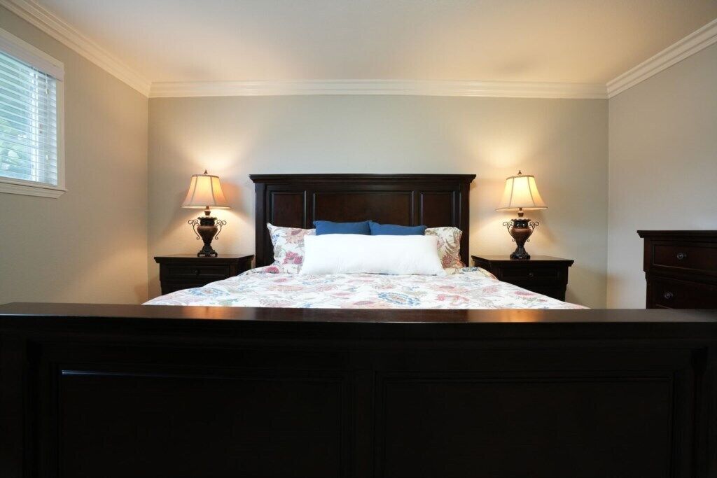 Une chambre à coucher avec une tête de lit en bois foncé, deux lampes de chevet, une couvre-lit à motifs floraux et une fenêtre avec des stores.