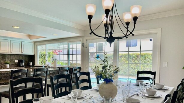 A dining area with a table set for six, surrounded by chairs, a chandelier, and a view of a pool through large windows. - Large Family Pool Home 2.5 Miles To Disney w/3 King Beds!!! (Anaheim)
