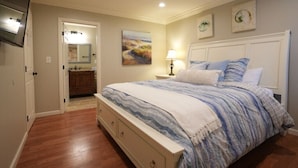 A bedroom with a bed, a nightstand, a lamp, a painting, and a bathroom visible through an open door. - Large Family Pool Home 2.5 Miles To Disney w/3 King Beds!!! (Anaheim)