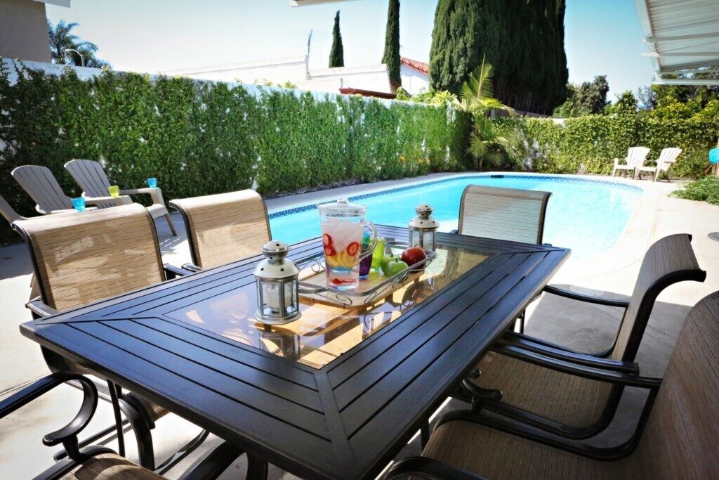 A glass-top table with chairs, a pool, and a hedge in the background.