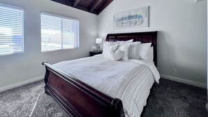 Un dormitorio con cama de madera, ropa de cama blanca, una mesita de noche con lámpara y un cuadro en la pared.