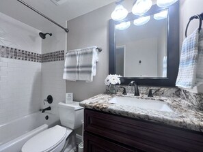 A bathroom with a white toilet, a bathtub, a sink with a marble countertop, and a mirror with lights.