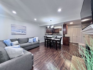 A modern living room with a gray sofa, dining table, and chairs, a kitchen area with wooden cabinets, and a large window. - Luxurious 2 Bedroom Condo Shortest Walk To Disney (Anaheim)