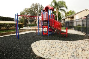 A playground with a red and blue play structure, surrounded by a paved area and a fence. - Luxurious 2 Bedroom Condo Shortest Walk To Disney (Anaheim)
