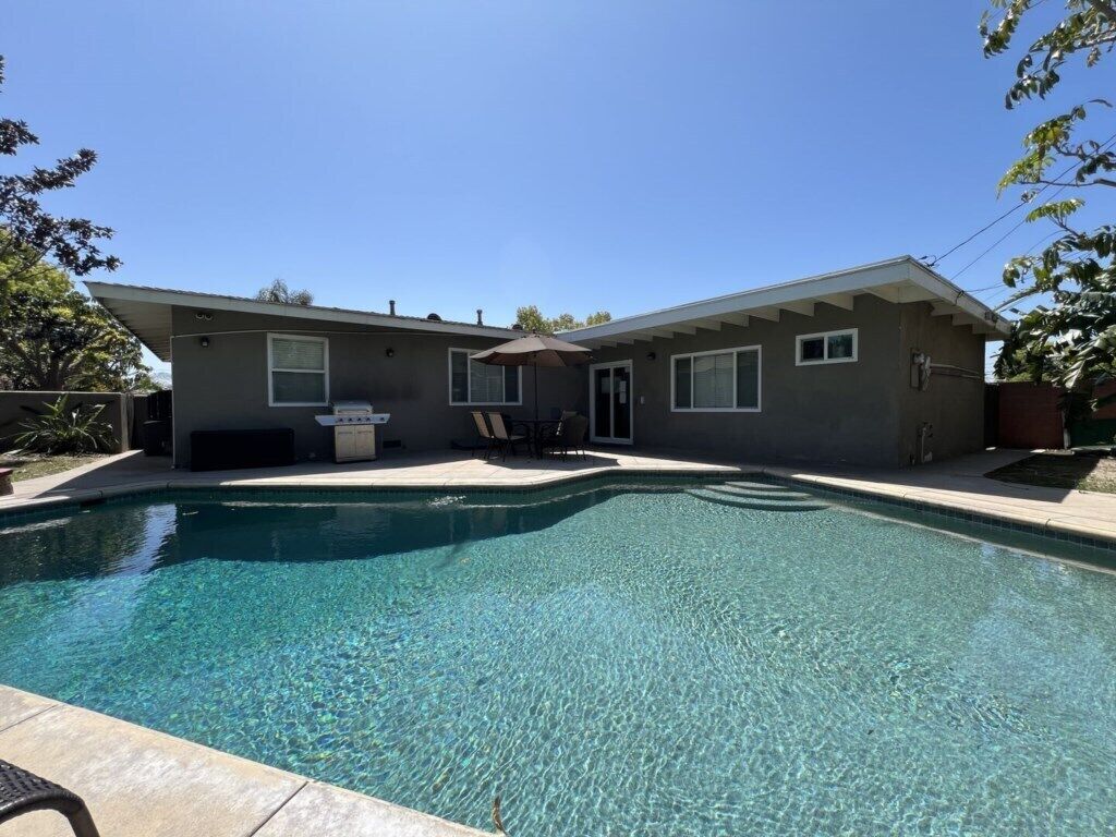 A house with a swimming pool, a patio with chairs, and a small outdoor kitchen.