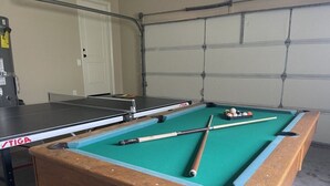 Una mesa de billar con mesa TIGA, tacos y bolas en una habitación con puerta de garaje y puerta blanca.