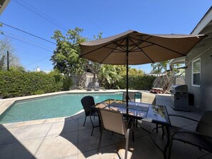 Een zwembad met een patio tafel en stoelen, een grote parasol en een barbecue.