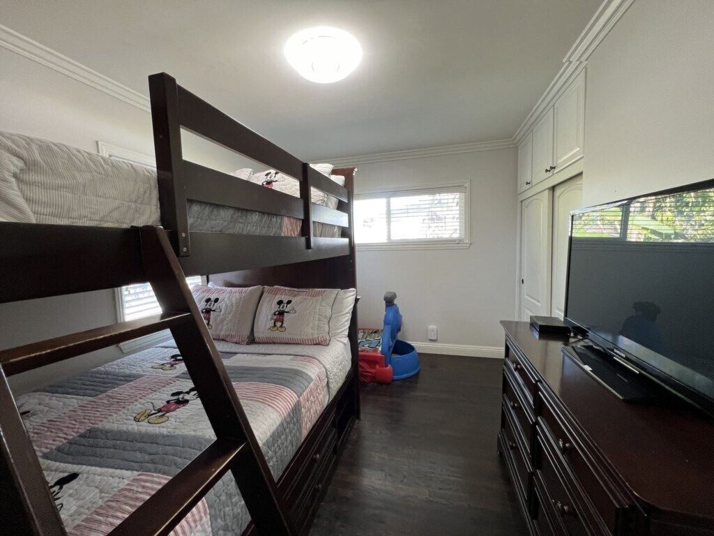 Een slaapkamer met een stapelbed, een televisie, een dressoir en een raam met jaloezieën.