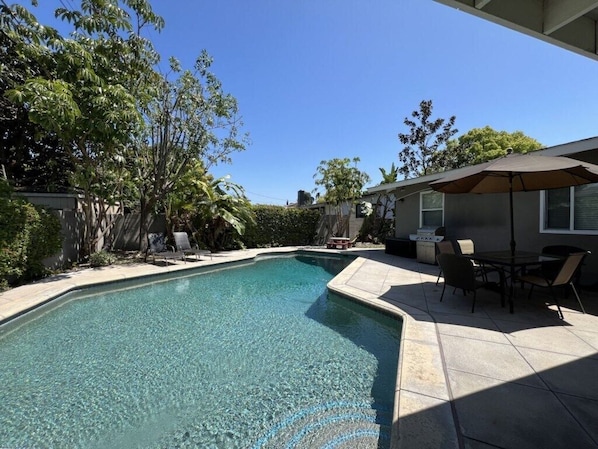 A swimming pool with clear water, surrounded by a patio area with chairs and a table, and a covered seating area. - Gorgeous Pool Home 1/2 Mile Walk to Disney (Anaheim)