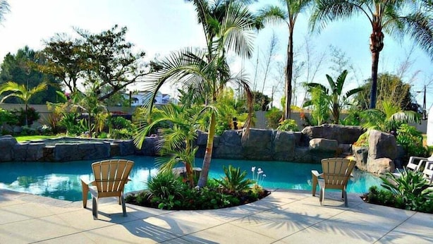 A pool area with two chairs, palm trees, and a rocky backdrop. - Incredible Estate 5 Bedrooms 3 Bathroom W/resort Style Rock Pool & Jacuzzi (Fullerton)