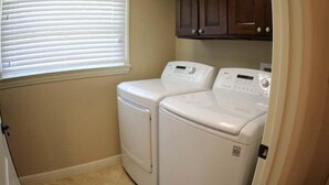 A laundry room with two white front-loading washers and dryers, beige walls, and a window with blinds. - Incredible Estate 5 Bedrooms 3 Bathroom W/resort Style Rock Pool & Jacuzzi (Fullerton)