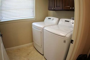 A laundry room with two white front-loading washers and dryers, beige walls, and a window with blinds. - Incredible Estate 5 Bedrooms 3 Bathroom W/resort Style Rock Pool & Jacuzzi (Fullerton)