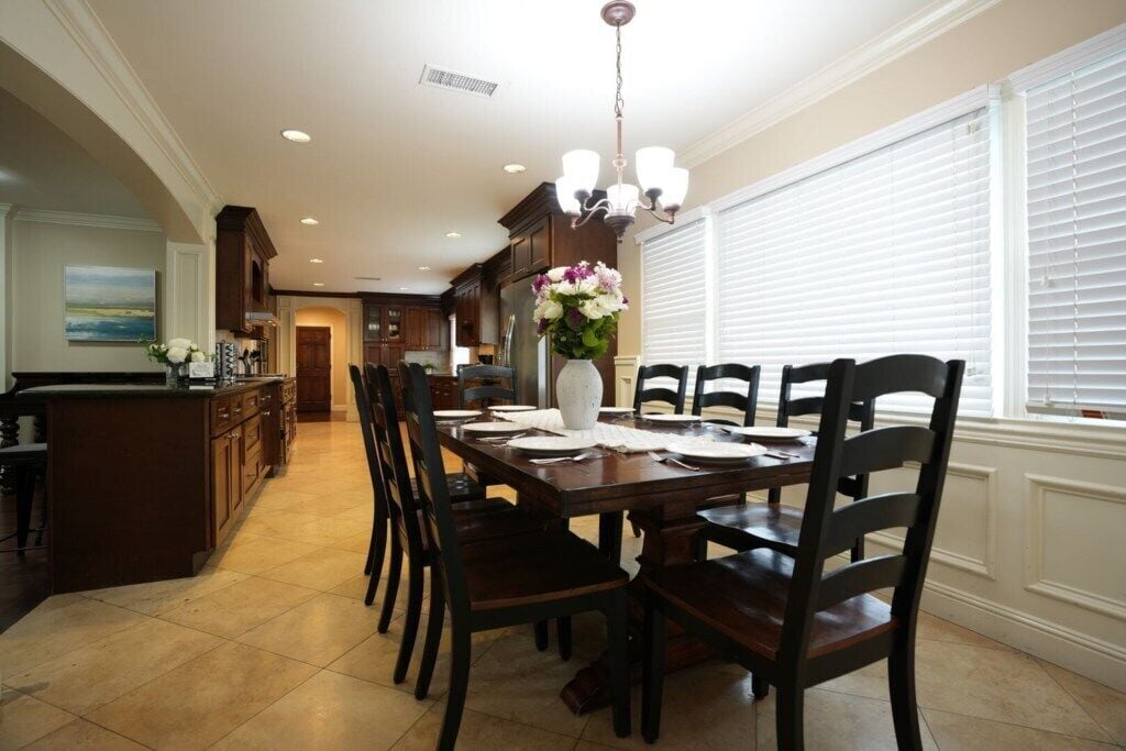 A dining area with a wooden table, dark chairs, a vase with flowers, and a kitchen in the background.