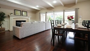 A living room with a fireplace, a sofa, a dining table with chairs, and a large window. - Incredible Estate 5 Bedrooms 3 Bathroom W/resort Style Rock Pool & Jacuzzi (Fullerton)