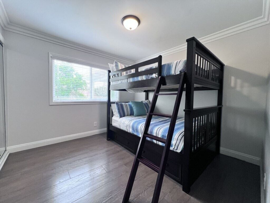 A bunk bed with a ladder, a window with blinds, and a ceiling light fixture.