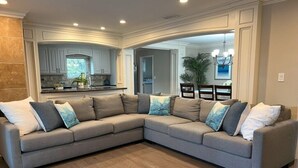 A modern living room with a large L-shaped sectional sofa, a kitchen in the background, and a dining area with a chandelier. - Fabulous 6 Bedroom , 4 Bath Pool W/jacuzzi Home (Orange)