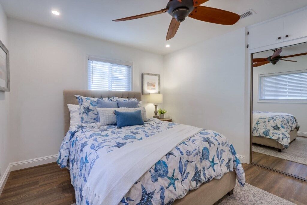 Un dormitorio con cama, ventilador de techo, lámpara de noche, ventana y espejo.