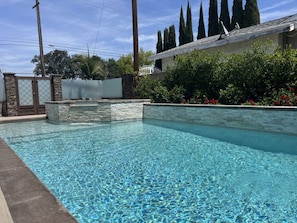 A clear swimming pool with a stone border, surrounded by a fence and greenery. - Beautiful Remodel 3 Bedroom 2 Bathroom Home With Pool & Jacuzzi (Fullerton)