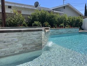A swimming pool with stone walls, surrounded by greenery and a house in the background. - Beautiful Remodel 3 Bedroom 2 Bathroom Home With Pool & Jacuzzi (Fullerton)