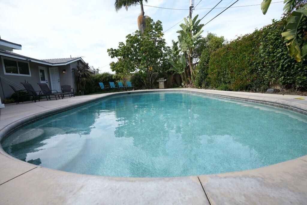 A swimming pool with a curved edge, surrounded by a patio with chairs, a house, and lush greenery.